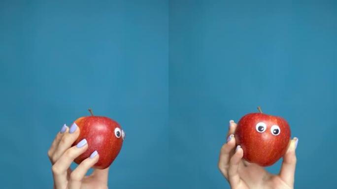 红苹果与眼睛在一个女人的手特写。苹果在蓝色背景上四处张望。垂直视频。
