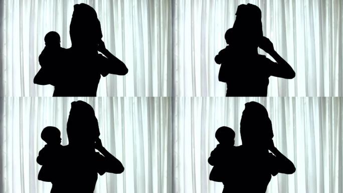 一个女人站在窗前抱着婴儿并在手机上聊天的剪影
