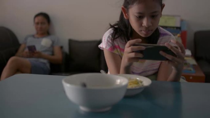 亚洲可爱女孩玩手机游戏，在家检疫时坐在远离母亲的地方。女性青少年在吃早餐时使用智能手机。