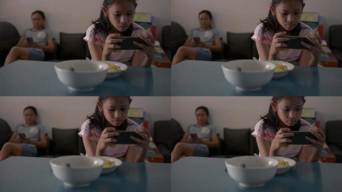 亚洲可爱女孩玩手机游戏，在家检疫时坐在远离母亲的地方。女性青少年在吃早餐时使用智能手机。