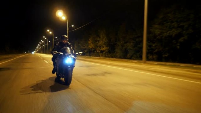 戴着头盔的年轻人在晚上的城市街道上快速骑现代运动摩托车。摩托车手在夜路上骑摩托车。开着前灯驾驶自行车