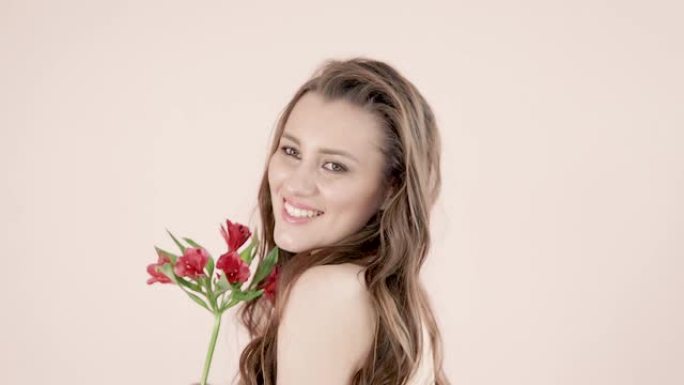一位年轻漂亮的女人，有着棕色的波浪长发，手里拿着一朵红色的花，温柔地微笑着，调皮地转身