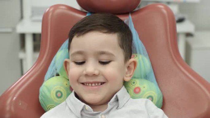 学龄前的男孩坐在舒适的牙科椅上，特写肖像，悲伤地看着相机，尴尬，开始微笑，放低眼睛，拿出玩具，在医生