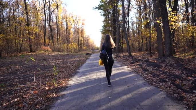 在公园散步的匿名女人。在安静的秋季公园里，年轻女性带着装饰性背包沿着小路行走的背景图