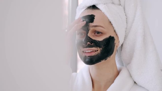 用毛巾包裹头发的漂亮女人正在脸上涂上黑色面具