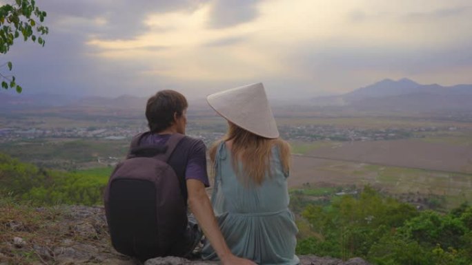 一对年轻的游客坐在山顶上，可以欣赏山谷的壮丽景色。她戴着一顶亚洲帽子