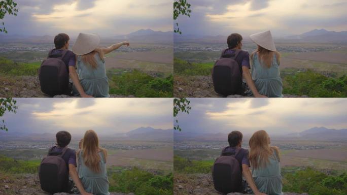 一对年轻的游客坐在山顶上，可以欣赏山谷的壮丽景色。她戴着一顶亚洲帽子