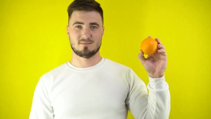一个年轻人手里拿着一个新鲜的橘子。一个穿着白色运动衫的男人眨眨眼，微笑着。适当营养的概念。黄色简单背
