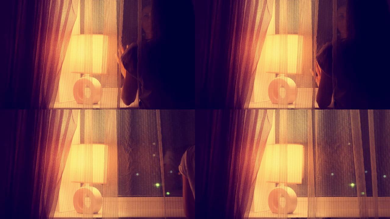 女人站在带窗帘和台灯的夜窗前。女性在窗前悲伤