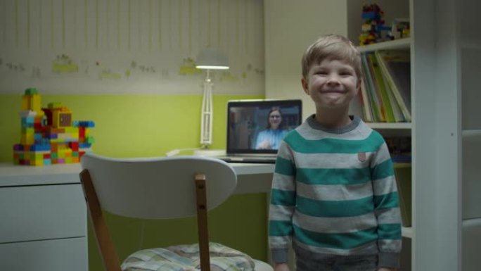 学龄前儿童男孩微笑着站在家里的儿童房，后面是笔记本电脑上的在线课程。电子学习快乐的孩子看着相机。