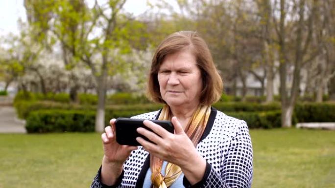春天，穿着蓝色毛衣和围巾的欧洲老年妇女的肖像，使用黑色智能手机在城市公园用手指点击触摸屏，背景是绿色