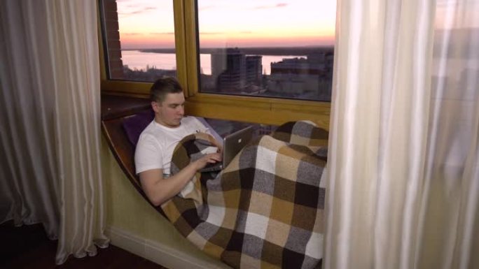 一个年轻人正在笔记本电脑上打字。一个人躺在窗边的窗台上，手里拿着笔记本电脑。晚上以外