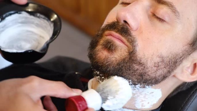 专业理发师在理发店将剃须泡沫涂在客户皮肤上。。年轻人在理发店把他未刮胡子的胡须剪成形状。男性美容理念