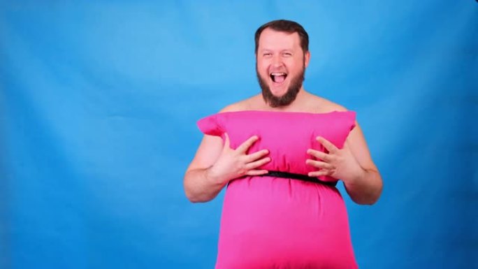 穿着粉红色枕头连衣裙的有趣的大胡子家伙在蓝色背景上抚摸着他的胸部。疯狂的检疫。有趣的打扫房子。时尚2