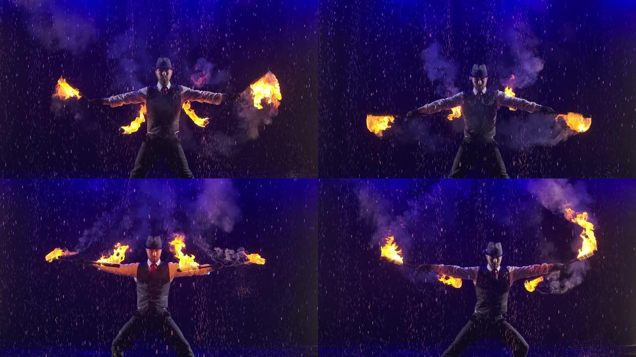 专业的马戏团表演者在一个黑烟的工作室和蓝光的背景下，在雨水下旋转燃烧的火炬。慢动作特写