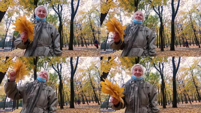 戴着防护面具和一束秋叶的老妇人在户外的秋季公园里玩得很开心。