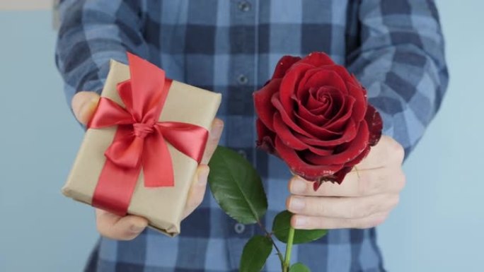 双手正在赠送带有红色蝴蝶结的包装礼物，朝相机移动，特写镜头。男人拿着小礼盒，手里拿着玫瑰去参加情人节