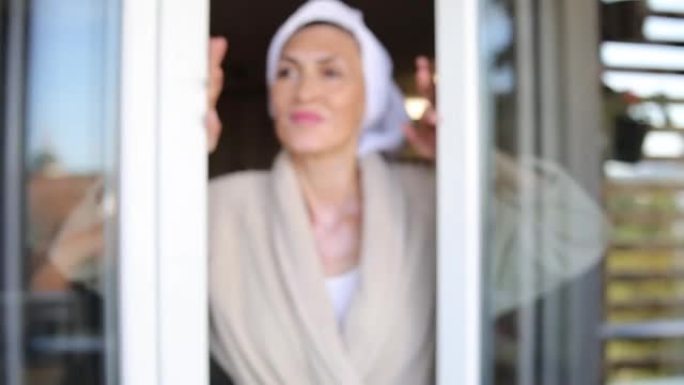 一个漂亮的老妇人打开阳台门，享受新鲜阳光明媚的早晨的慢动作镜头
