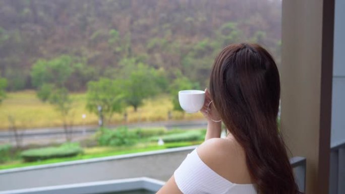 亚洲妇女喝咖啡或茶，在公寓阳台上眺望山景