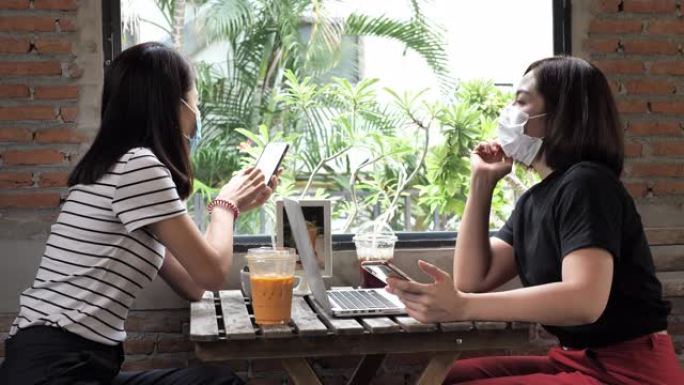 两个亚洲女性最好的朋友戴着医疗脸马克，喜欢在舒适的咖啡店里用笑脸见面交谈
