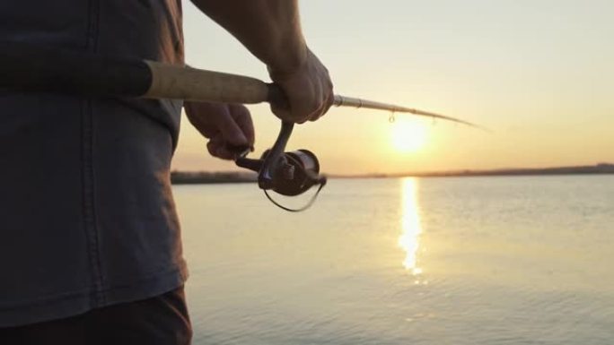 用钓鱼竿的卷轴处理旋转，以对抗橙色日落慢动作。男子业余爱好在海上钓鱼收紧了鱼线卷轴的夏天。镜头光斑。