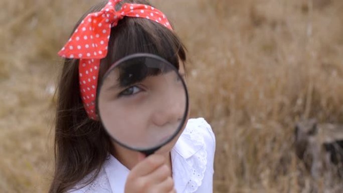 探索世界的孩子。特写四岁好奇的蓝眼睛小女孩穿着白色上衣和圆点红领巾的肖像。透过放大镜看着相机。4k 
