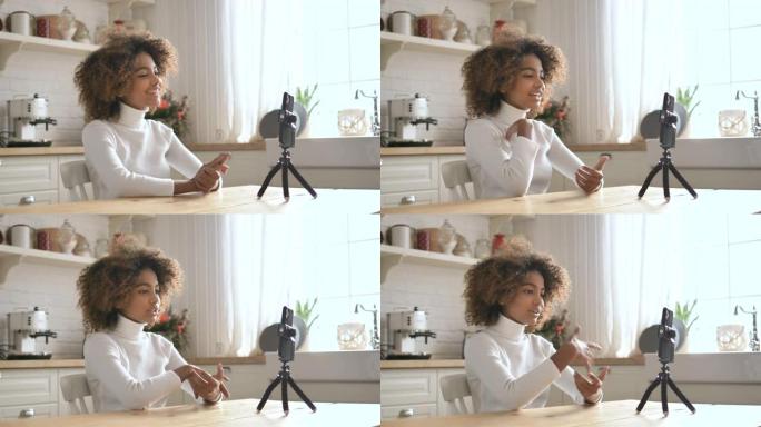 可爱的社交非洲女孩博客作者坐在家里的厨房里用手机写她的视频博客