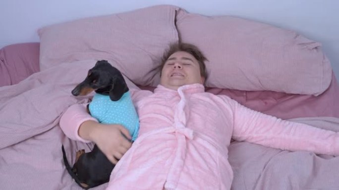 穿着有趣的粉红色睡衣和腰带的年轻快乐男人在拥抱他心爱的腊肠狗漫长的辛苦一天后，躺在床上准备睡觉和放松