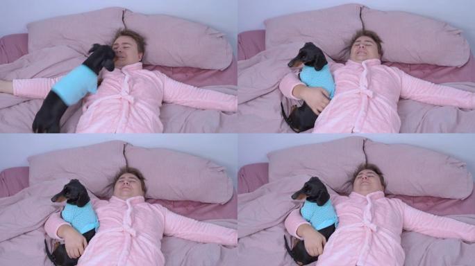 穿着有趣的粉红色睡衣和腰带的年轻快乐男人在拥抱他心爱的腊肠狗漫长的辛苦一天后，躺在床上准备睡觉和放松