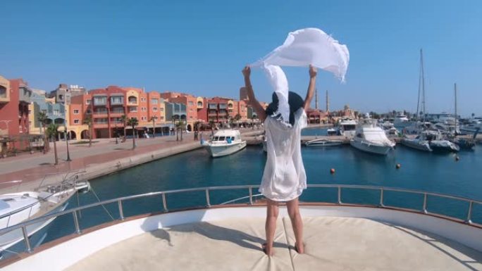 穿着白色连衣裙和帽子挥舞着织物围巾的时尚模特站在埃及赫尔加达码头的豪华游艇上