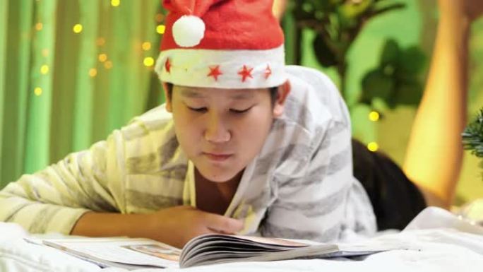 快乐的亚洲男孩戴着圣诞老人的帽子，看书，晚上躺在床上，穿着圣诞装饰品，生活方式的概念。