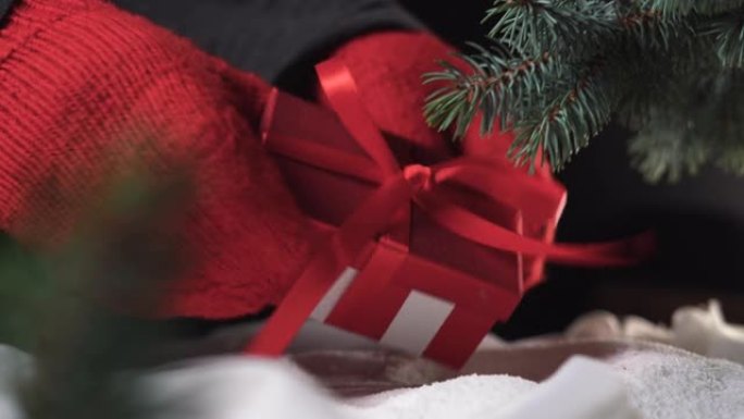 一个带有蝴蝶结的红色盒子躺在云杉树枝下的雪地里。手套中的两只手拿着礼物