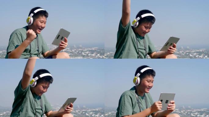 快乐的亚洲男孩戴着耳机，在阳光明媚的日子里用数字平板电脑在山顶上，有自然背景。