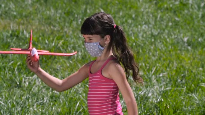 特写镜头，可爱的小女孩戴着口罩，在绿色草坪上玩红色玩具飞机。大流行后恢复安全飞行。4K 50fps慢