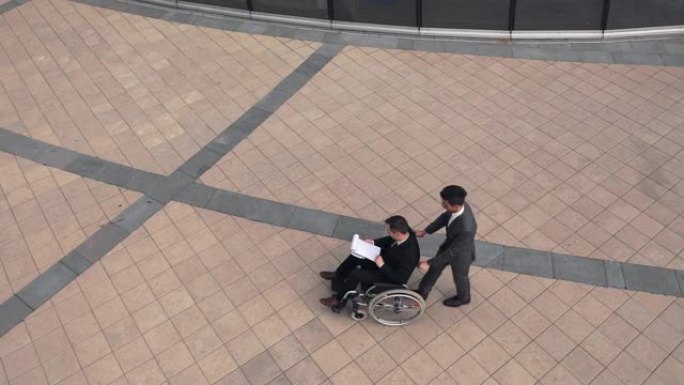两个商人在公司大楼附近的户外聊天。同事坐在轮椅上。鸟瞰图。从顶部看。