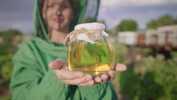 带蜂蜜的养蜂人。一位女农民从养蜂场展示蜂蜜。玻璃罐子里的黄色蜂蜜