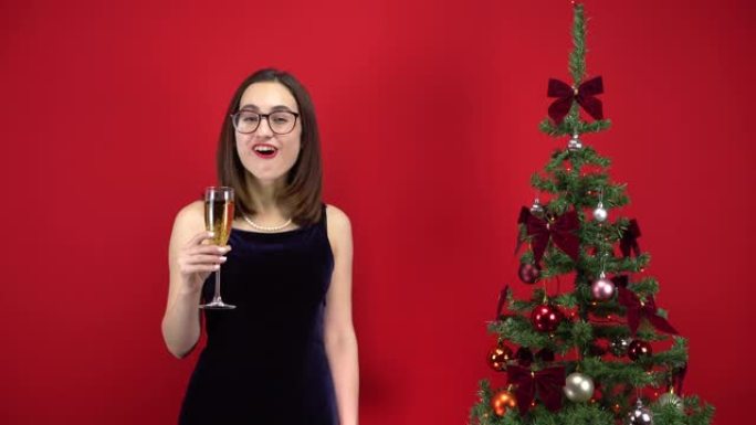 年轻女子在红色背景的圣诞树附近喝了一杯香槟，度过了假期的五秒钟。一个戴着眼镜和晚礼服的女孩。