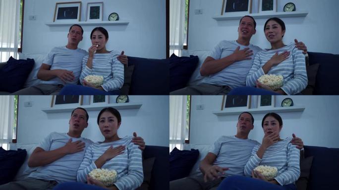 30多岁的年轻成年丈夫和妻子坐在家里的沙发上，在电视上观看恐怖娱乐节目的肖像。亚洲害怕和害怕的夫妇在