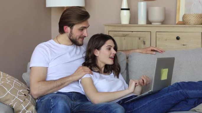 幸福的夫妻坐在客厅的沙发上放松，一起使用笔记本电脑在线购物，看电影，喝红酒。
