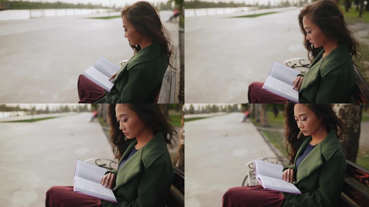 年轻的亚洲妇女坐在河边的长凳上读书