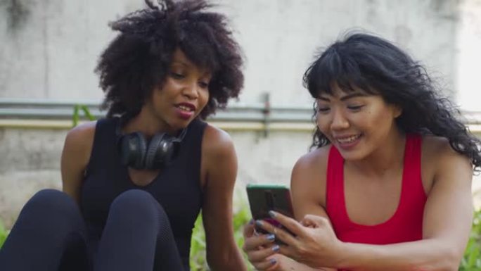 由两个女性健身朋友组成的多元化团体在公园里聊天和使用手机。年轻女性在户外休息和微笑
