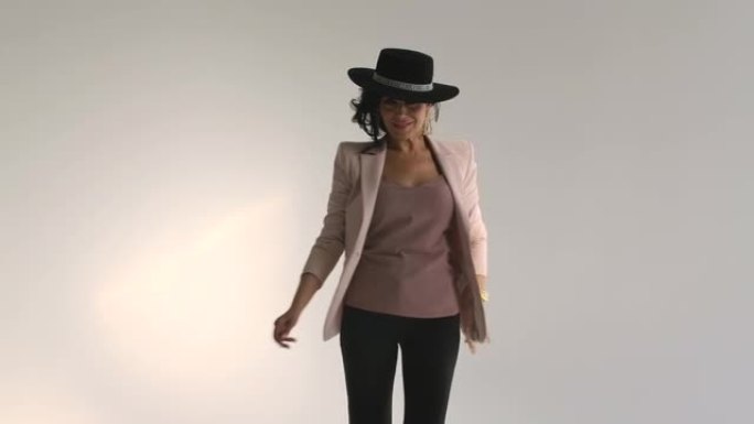 性感的女人在黑色裤子和一顶帽子摆姿势在一个灯光工作室对白墙的背景。可爱的黑发微笑和展示服装。关闭了。