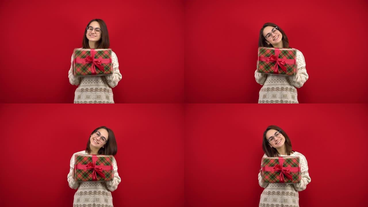 一位戴着眼镜的年轻女子手里握着圣诞礼物，欢欣鼓舞。在红色背景的摄影棚里拍摄。