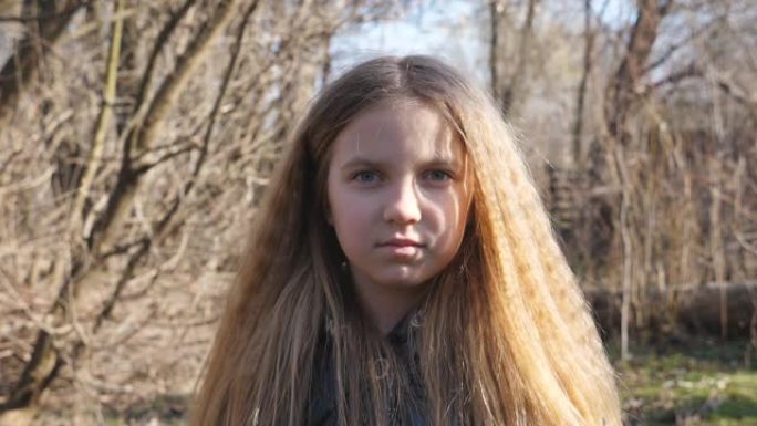 不快乐的金发女孩在户外看着相机。早春森林模糊背景下的小孩肖像。脸上表情严肃的女性孩子的情绪。