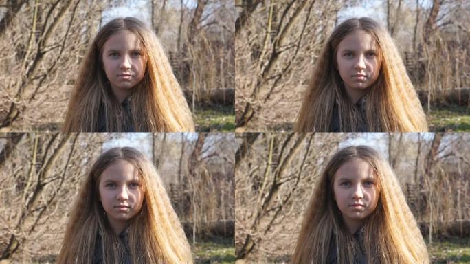 不快乐的金发女孩在户外看着相机。早春森林模糊背景下的小孩肖像。脸上表情严肃的女性孩子的情绪。