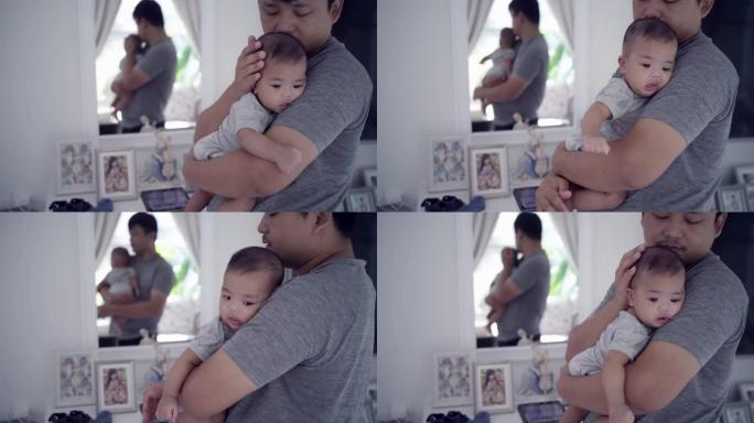 一位年轻的亚洲全职父亲抱着并安抚婴儿，在爱与关怀的陪伴下在客厅睡觉。