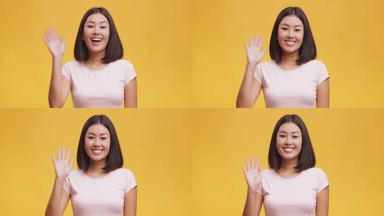善于交际的年轻亚洲女性在镜头前友好微笑，挥手示意你好，黄色背景