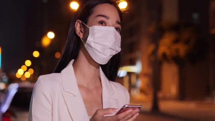 商业亚洲妇女在使用智能手机时，戴着防护口罩站着等待汽车在高峰时段接车到城市。