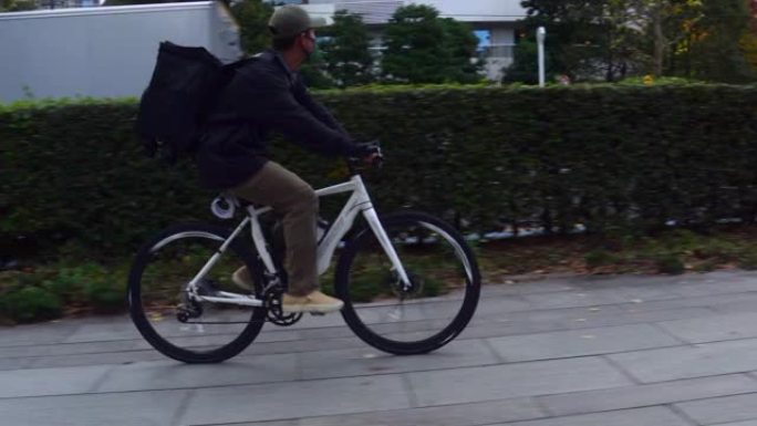 带着黑色背包和防护面具的年轻人快递员在街上骑着自行车穿过城市，运送食物。送餐概念。