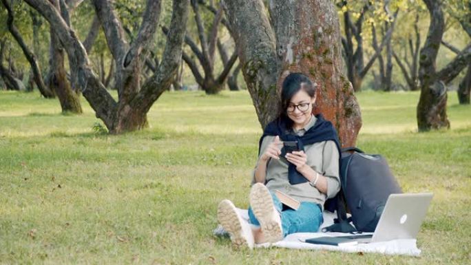 戴着眼镜微笑的年轻女子坐在公园的草地上滚动社交网络中的新闻提要